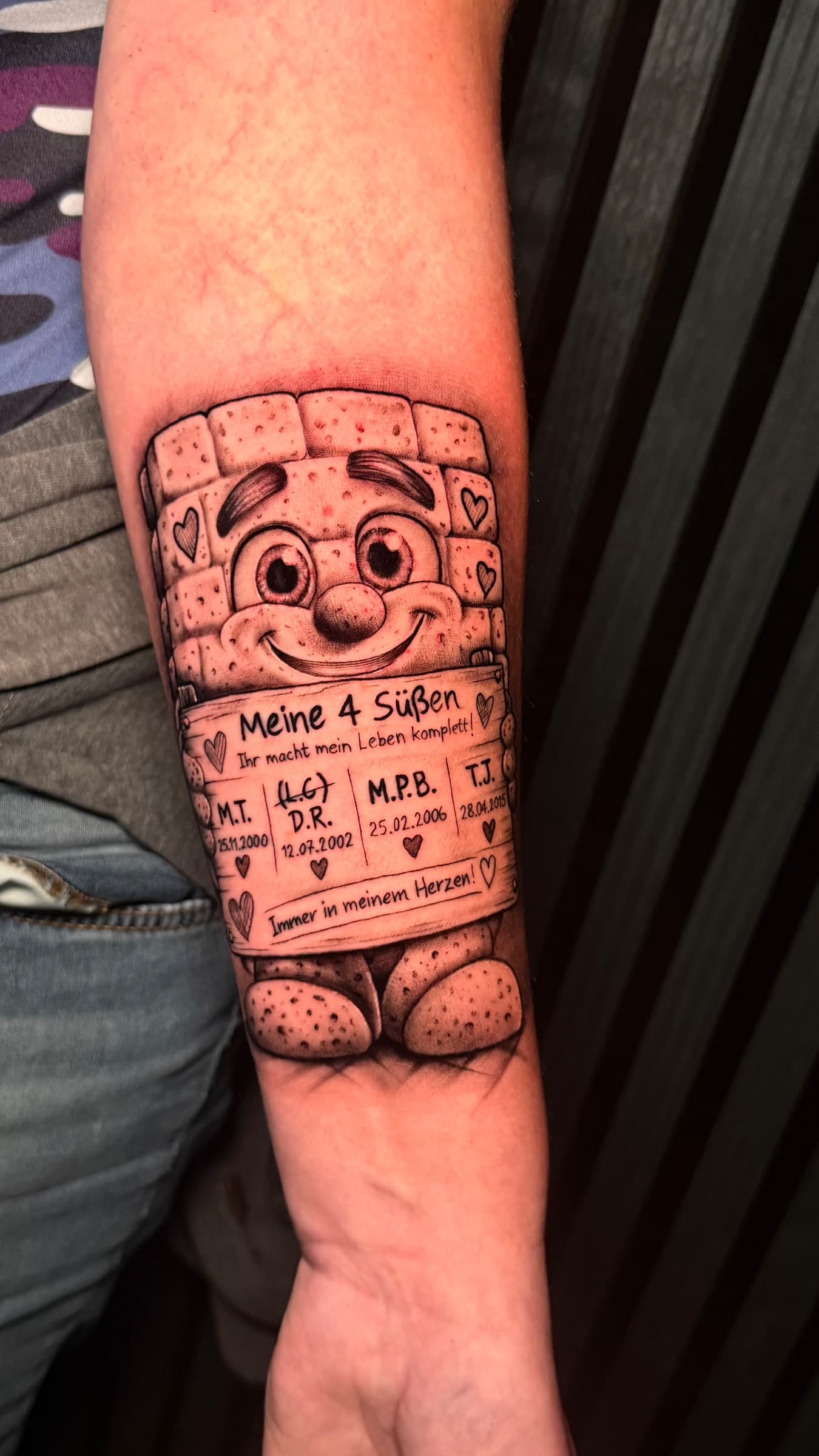 Ein persönliches Black-&-Grey-Tattoo einer niedlichen Mauer-Figur mit den Namen und Geburtsdaten ihrer vier Kinder sowie kleinen Herzen.